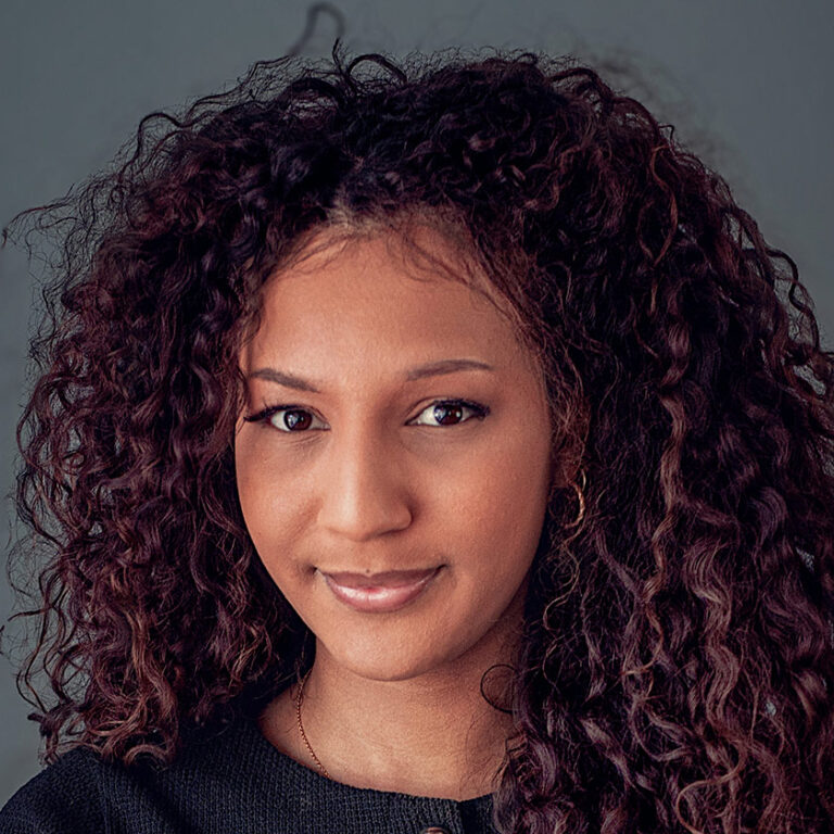 Junge Frau mit lockigem Haar, lächelt sanft, trägt dunklen Pullover vor grauem Hintergrund.
