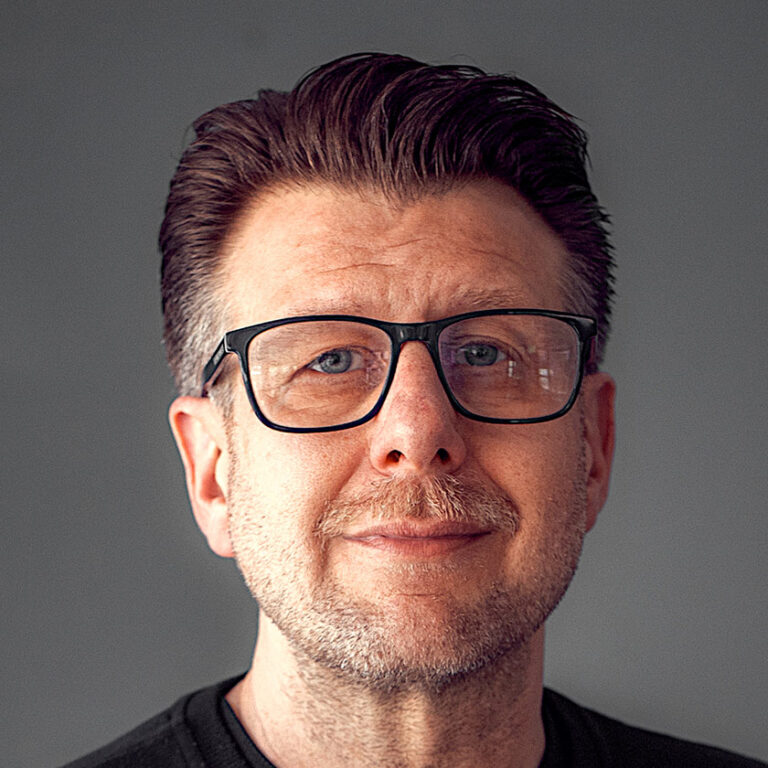 Mann mit Brille und leichtem Lächeln vor grauem Hintergrund. Kurze Haare, braune Haare.
