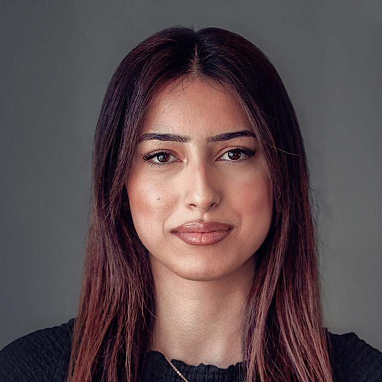 Person mit langen Haaren, neutralem Gesichtsausdruck, schwarzes Oberteil vor grauem Hintergrund.